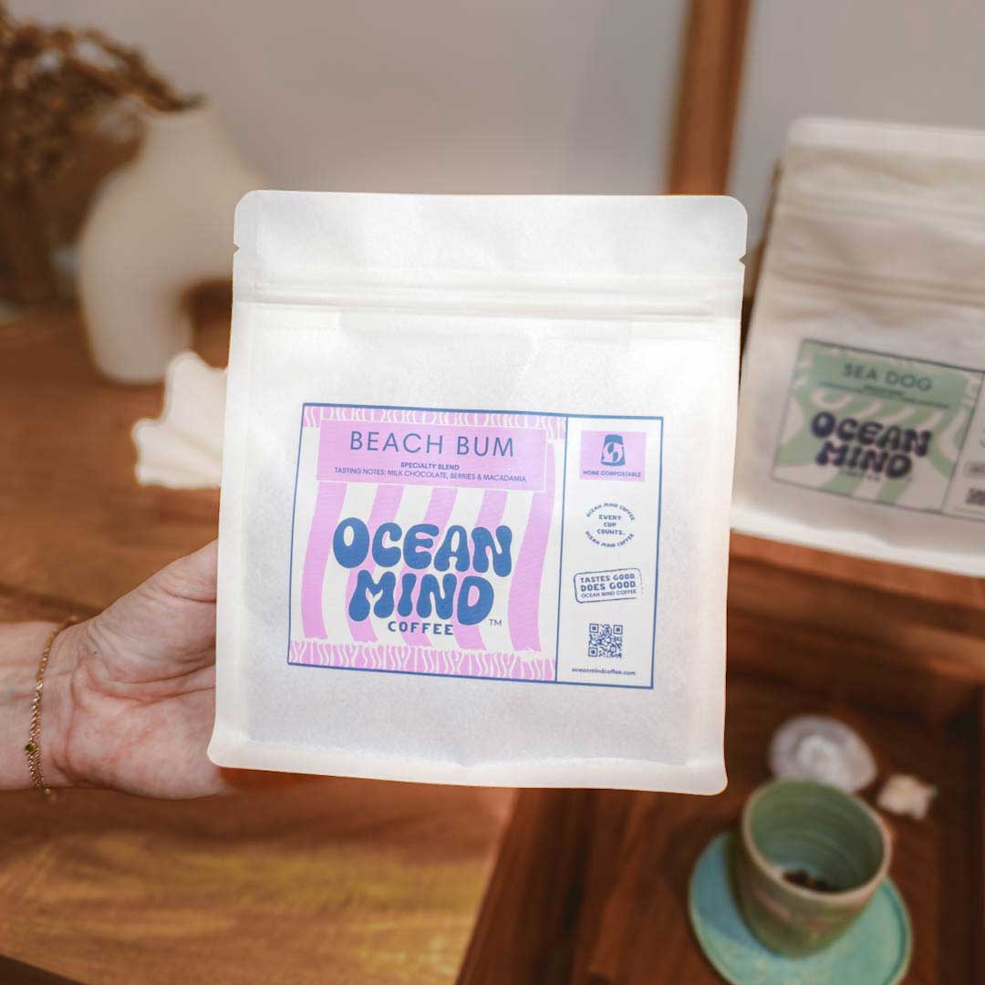 Hand holding a package of Ocean Mind Coffee labeled 'Beach Bum' on a wooden table