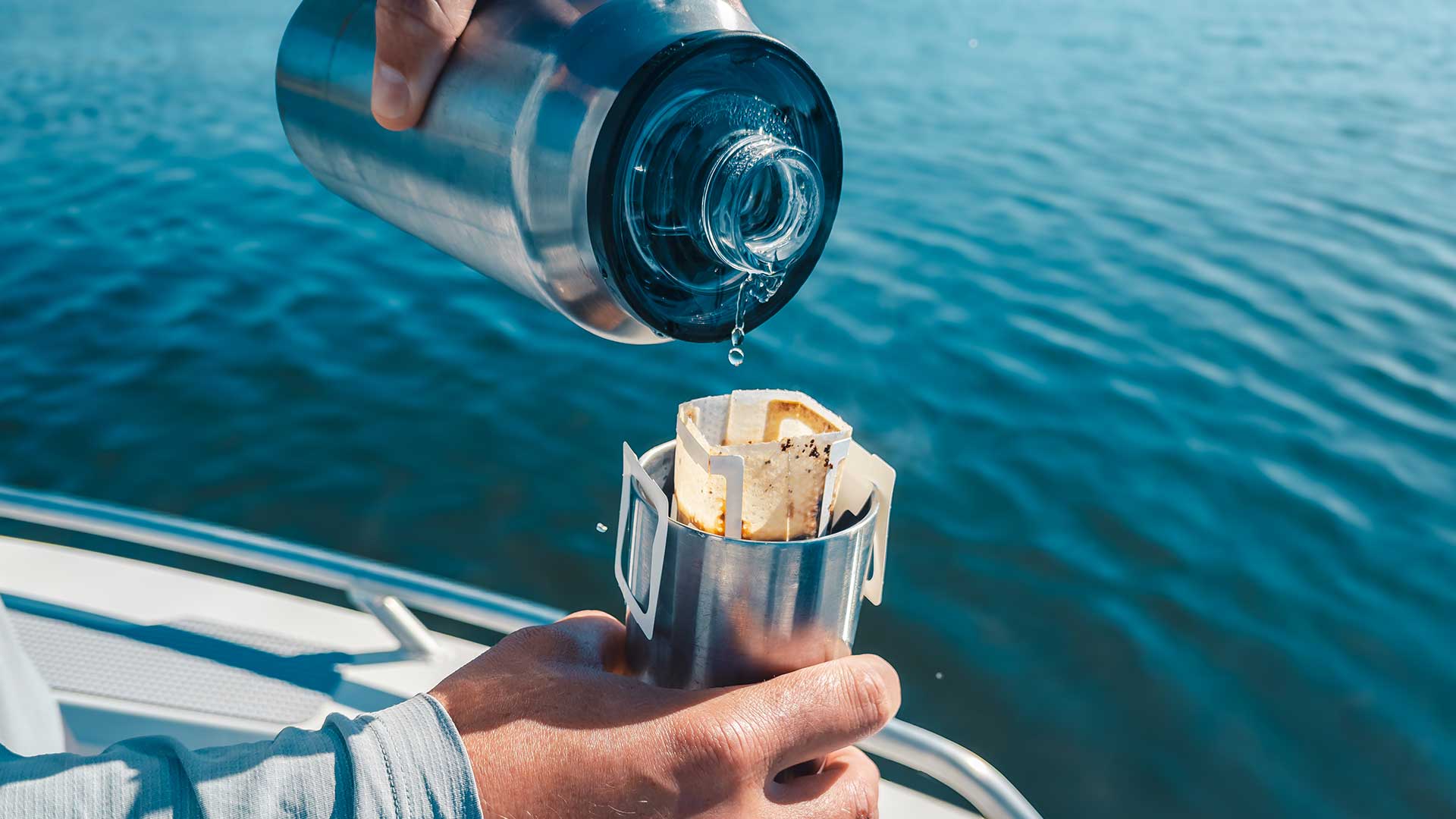 Person pouring hot water from a thermos into a coffee filter over a body of water.