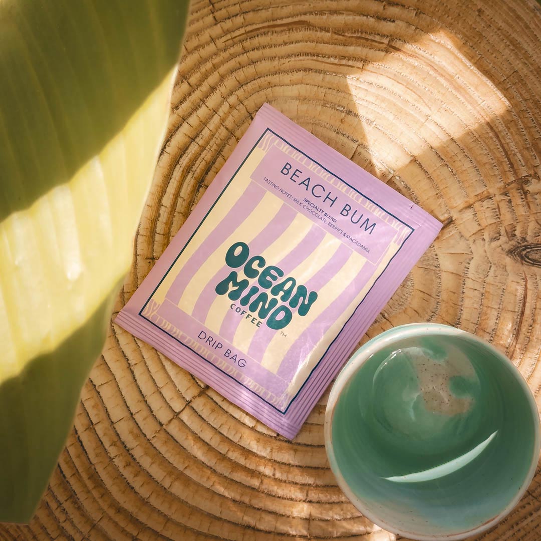 Beach Bum Ocean Mind coffee package on a woven surface with a green cup.