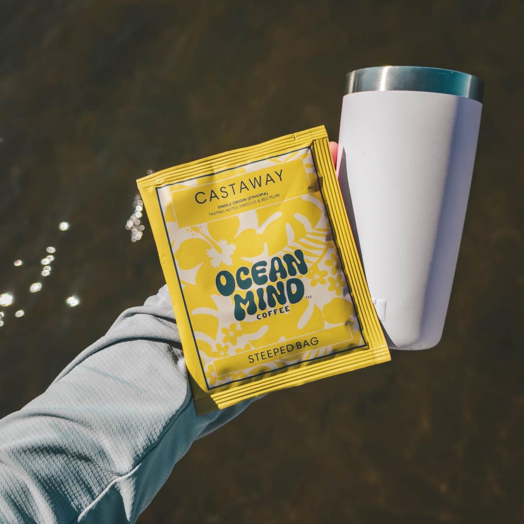 Castaway Ocean Mind Coffee steeped bag held by a hand with a white cup in the background