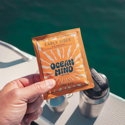 Hand holding an Early Riser Ocean Mind Coffee drip bag with a scenic background
